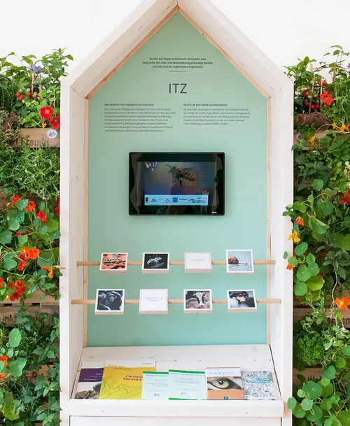 Ausstellungsstand in Form eines kleinen Holzhäuschens mit einer mintgrünen Rückwand, der Überschrift „ITZ“, einem Bildschirm, kleinen Bildtafeln und Informationsmaterial, umgeben von grünen Pflanzen mit orangefarbenen Blüten.