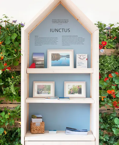Ausstellungsstand in Form eines kleinen Holzhäuschens mit einer blauen Rückwand, der Überschrift „IUNCTUS“, mehreren Regalen mit gerahmten Bildern, Büchern und Informationsmaterial, umgeben von grünen Pflanzen mit orangefarbenen Blüten.