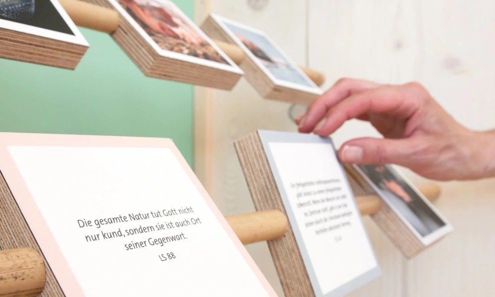 Nahaufnahme einer Hand, die eine kleine Tafel mit Text von einem Holzdisplay nimmt, umgeben von weiteren Tafeln mit Zitaten und Bildern.