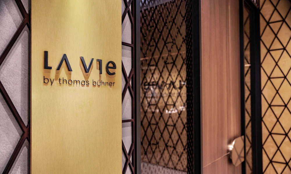 Logo mit Schriftzug 'LA VIE by thomas bühner' auf einer goldenen Wand, daneben eine Glasfront mit gitterartigem Muster.