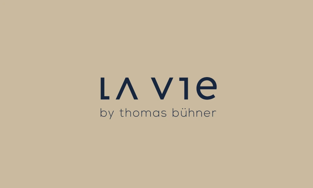 Logo von 'La Vie by Thomas Bühner' in dunkelblauer Schrift auf beigefarbenem Hintergrund.