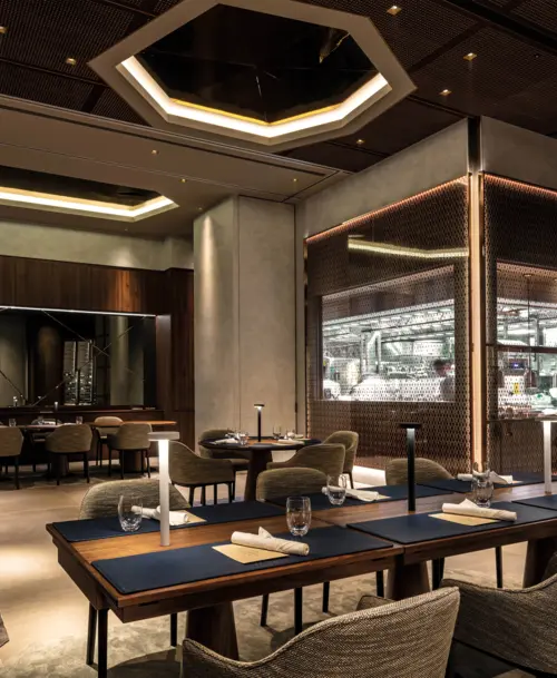 Elegant gestaltetes Restaurantinterieur mit moderner Beleuchtung, Holztischen, die mit Servietten und Gläsern gedeckt sind, und einer offenen Küche, die durch eine Gitterwand im Hintergrund abgetrennt ist.