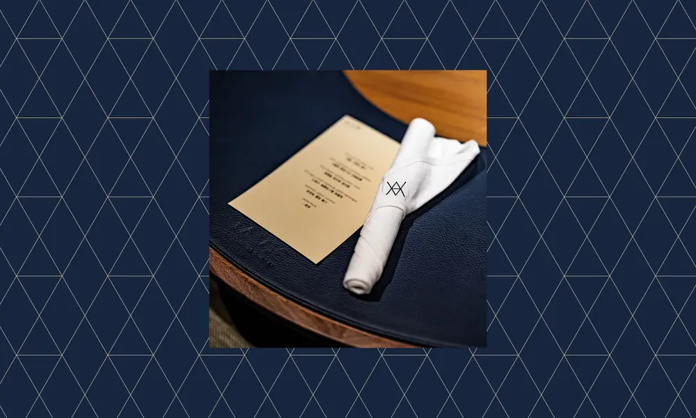 Elegant gedeckter Tisch mit beiger Menükarte und weißer, gerollter Serviette mit minimalistischem Logo auf dunkelblauer Tischdecke