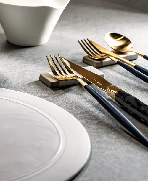 Elegantes Besteck mit goldenen Details und dunklen Griffen, platziert auf minimalistischen Haltern, daneben ein weißer Teller und eine Schale auf grauer Oberfläche.