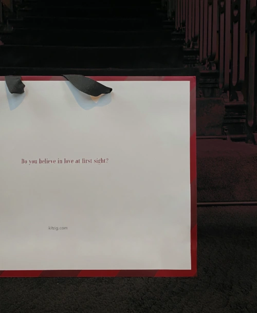 Weiße Einkaufstasche mit rotem Rand und schwarzem Henkel, bedruckt mit der Frage „Do you believe in love at first sight?“, auf einer dunklen Treppe platziert.