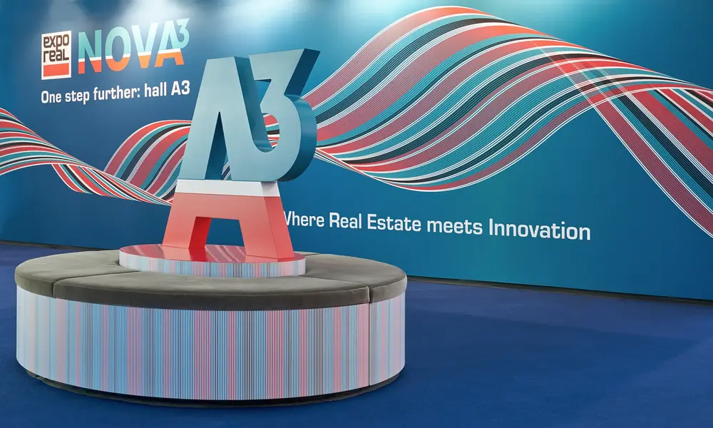 Messestand mit einer großen "A3"-Skulptur auf einer runden Sitzbank, vor einer Wand mit dem Schriftzug "expo real NOVA3" und bunten Wellenmustern.