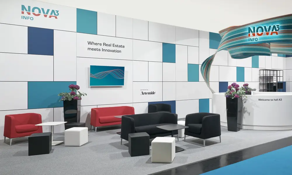 Moderner Messestand "NOVA³ INFO" mit einer geometrisch gestalteten weißen Wand, blauen Akzenten, Sitzmöbeln in Schwarz, Rot und Weiß sowie einem Empfangstresen mit Dekoration.
