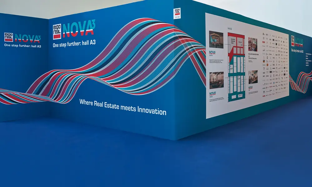 Messestand "expo real NOVA³" mit blauer Wand, bunten Wellenmustern und Informationsgrafiken, begleitet vom Slogan "Where Real Estate meets Innovation".