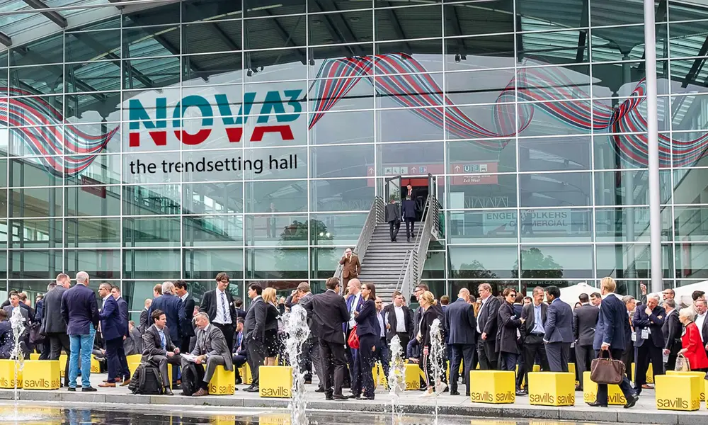 Menschenmenge in Business-Kleidung vor dem Eingang einer Messehalle mit der Aufschrift "NOVA³ the trendsetting hall", vielen gelben Taschen mit "savills"-Logo und moderner Glasfassade.