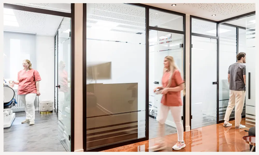 Moderner Innenbereich einer Zahnarztpraxis mit Glaswänden, behandelndem Personal und Patienten in einem hellen, minimalistischen Design.