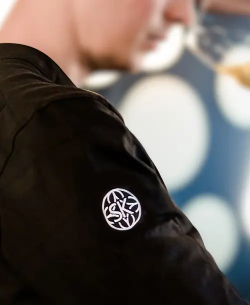 Mitarbeiter in schwarzer Uniform mit gesticktem Logo des „Hotel Stadt Kassel“ auf dem Ärmel, unscharfer Hintergrund mit stilvollen Lichtreflexionen.