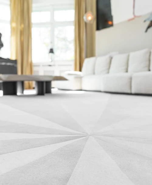 Helles Wohnzimmer mit cremefarbenem Designteppich und modernem Sofa vor großen Fenstern.