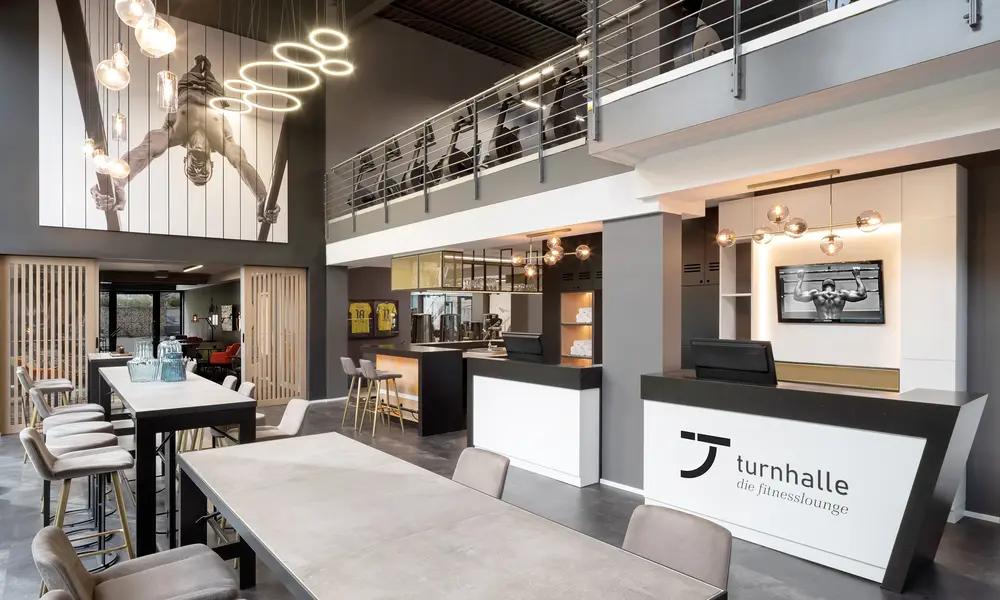 Moderner Empfangsbereich der „Turnhalle – Die Fitnesslounge“ mit stilvoller Einrichtung, hohen Decken und Fitnessmotiven an den Wänden.