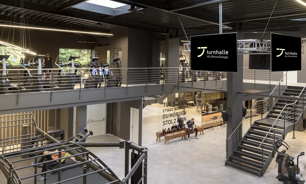 Modernes Fitnessstudio „Turnhalle – Die Fitnesslounge“ mit Trainingsgeräten, Empore, stilvoller Einrichtung und Motivationsspruch an der Wand.