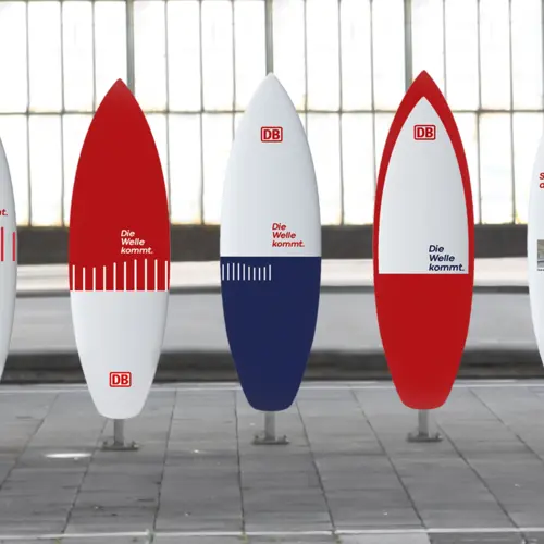 Mehrere surfbrettförmige Werbeaufsteller in Rot, Weiß und Blau mit dem DB-Logo und dem Slogan "Die Welle kommt." in einer modernen, industriellen Umgebung.