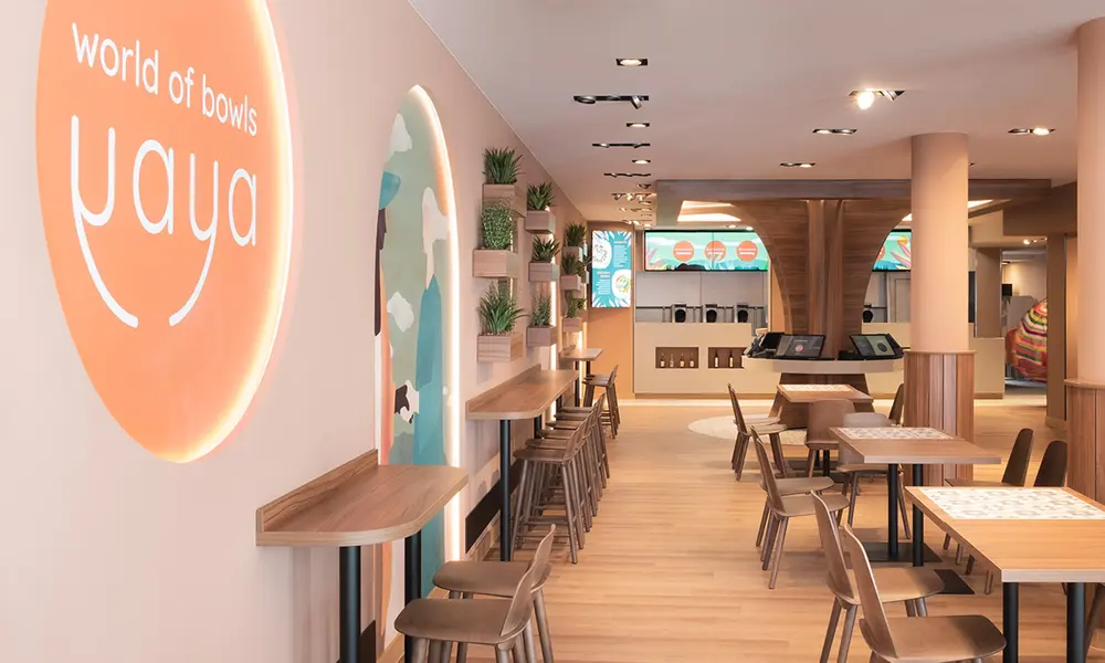 Innenraum des Gastro-Konzepts 'Yaya - World of Bowls' mit moderner Einrichtung, hellen Holzmöbeln und beleuchtetem Logo an der Wand.