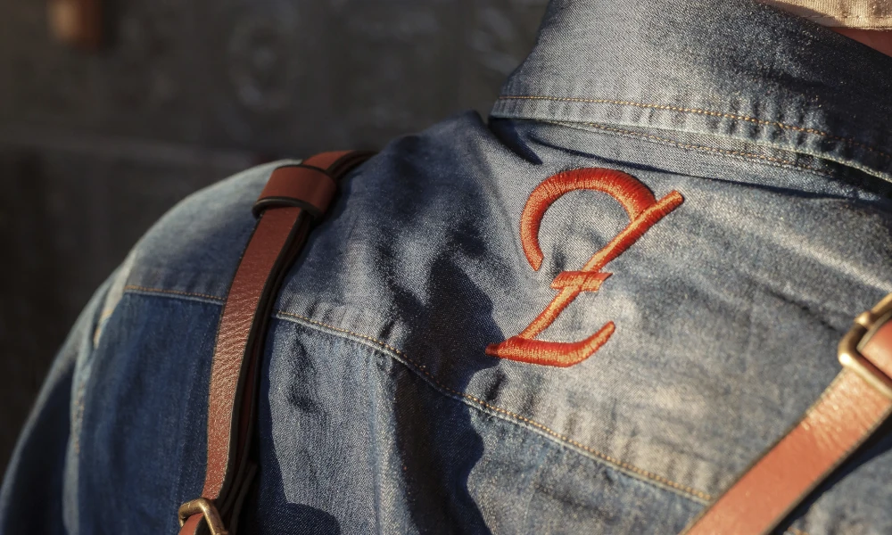 Gesticktes Zenz-Logo auf Jeanshemd mit Lederriemen – handwerklich geprägte Arbeitskleidung im Wirtshausstil, vereint Tradition, Authentizität und modernes Gastrodesign.